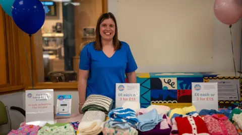 Grand Appeal Emma Stuart wearing blue nurse scrubs. She smiles as she stands in front of one of her fundraising stalls.