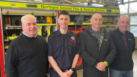 Four men of varying ages standing in front of a fire engine looking towards the camera, all wearing dark-coloured clothes. A family resemblance is visible between them.