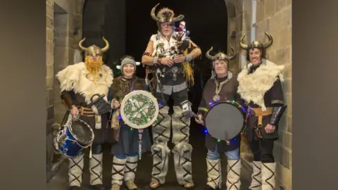 Supplied Five of the Vikings stand in the hallways of an old stone building. They are wearing various Viking accessories including helmets with horns, fur leggings and leather arm braces. The man in middle is on stilts, but dressed in a way that makes him look like a very tall Viking. There are holding various instruments including horns, pipes and drums.