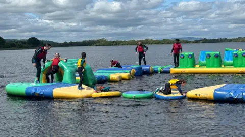 An inflatable playground sits on top of a lake, It's brightly coloured and has a series of obstacles. 8 people are on top of it, some standing, some falling. The people are dressed in wetsuits. The lake is surrounded by trees and there is a bright, but cloudy sky above. 