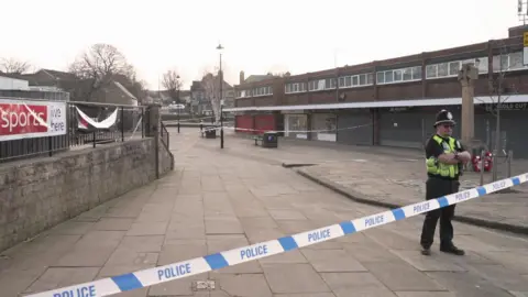 A cordoned‑off town centre area with blue and white police tape, a police officer standing guard, closed shopfronts with shutters down, a stone monument and benches nearby.