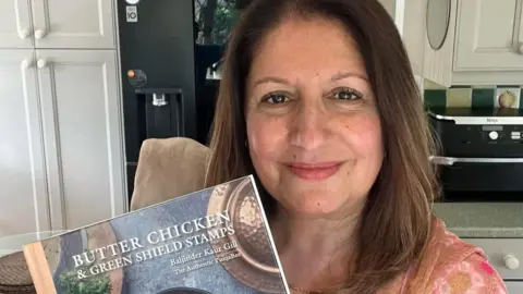 Baljinder Kaur Sangha-Gill Baljinder holding her cook book and smiling at the camera. She has mid-length brown hair and is standing in a kitchen.