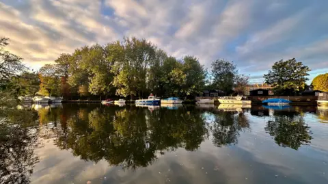 Emma H Streaks of sunlit cloud appear to spiral outwards from a cluster of trees beside a river with moored boats. The trees and cloud pattern are mirrored in the still water.