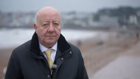 A smartly dressed man standing outdoors. He is wearing a black jacket with a shirt and tie underneath. He appears to be standing near a beach. The background is blurred.