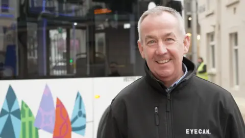 Mark Coxshall standing in front of a new LibertyBus bus. He's wearing a black jacket, with the charity's name 'Eyecan' written in white. He's smiling and has grey coloured hair. 
