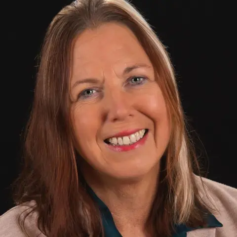 Claire Gallery-Strong Claire Gallery-Strong is smiling, she had long brown hair. She has a beige jacket and blue blouse on.