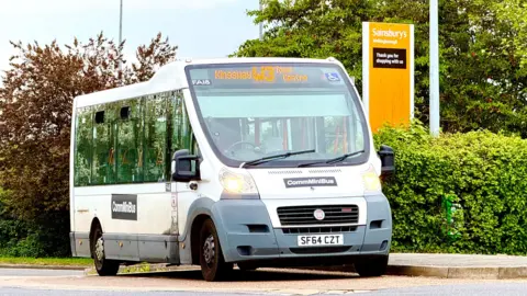 North Northamptonshire Council A white bus, by a Sainsbury's sign, it has writing at the top, and has a grey bumper. The bus is empty. Foliage is behind the bus and around a supermarket sign. 