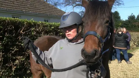 Paula Oakley stands next to her horse as she gives a TV interview. She is wearing equestrian gear. Behind her is a hedge, a hedge and a group of three people.