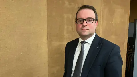 Amy Holmes/BBC Henry Vann in a dark suit and glasses stands indoors against a textured brown wall, with stacked chairs visible in the background.