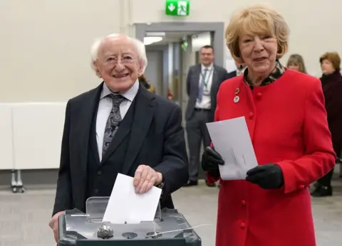 PA Michael D Higgins in a black suit and white shirt, wife Sabina in a red coat and black gloves, holding ballot papers and voting