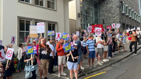 A large group of protestors outside of Guernsey's Royal Court. Most are holding placards on thin pieces of wood which say no to GST.