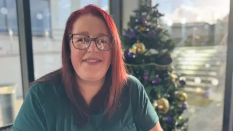 A women with long red hair smiling at the camera. She has patterned glasses on. She is wearing a dark green t-shirt. There is a Christmas tree behind her left hand side.
