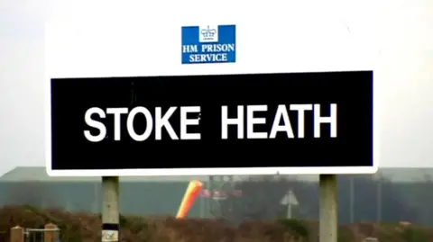 A black sign with the words Stoke Heath in white lettering. Behind them is landscape and a hedge
