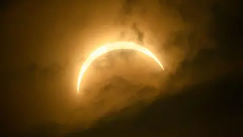 John O'Neill The eclipsed Sun forms a wider, glowing crescent surrounded by warm orange and gold tones. Thick clouds diffuse the sunlight, creating a striking halo effect around the crescent.