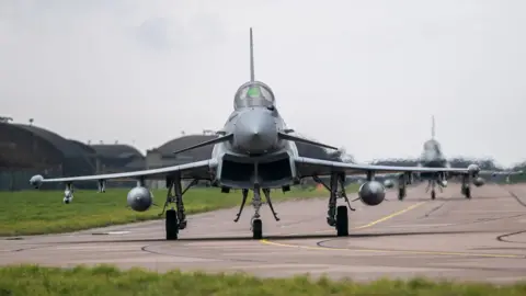 Getty Images The front of a taxiing Typhoon jet on the runway at RAF Coningsby in Lincolnshire with a second plane and hangars and a grassy area in the background. 