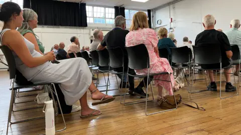 Three rows of people are pictured sitting on black chairs with steel metal frames in a community hall. They are all facing someone at the front of the hall.