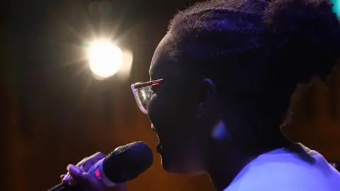 Naledi is a young singer standing at a microphone, in partial silhouette, with ultraviolet light causing her tee shirt to appear purple. She has her black curly hair tied back, and she wears glasses, and is singing into a microphone held in her right hand. A spotlight and rays of diffraction appear in the centre left of the image.