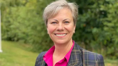 A smiling grey-haired woman standing on a lawn and in front of bushes, wearing a tartaned blazer and pink shirt.