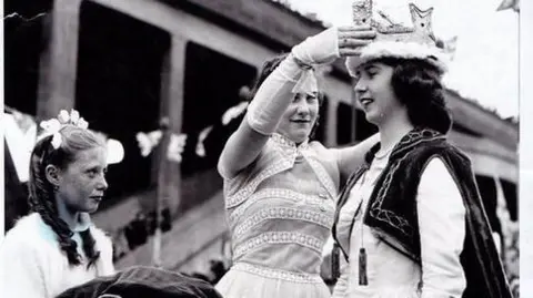 A black-and-white image of a woman being crowned by another woman. She is wearing a black cape and white dress; the woman placing the crown on her head is wearing a white dress. A young girl stands by with a cushion from which the crown has just been lifted - she is also wearing a white dress.