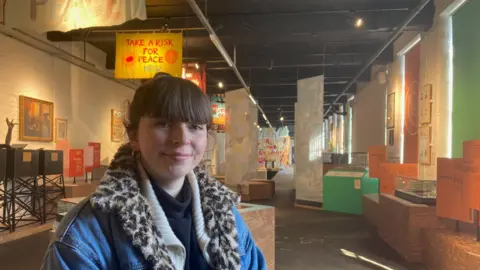 A woman wearing a faux leopard fur lined denim jacket standing in a long room with exhibits behind and to the side of her. 