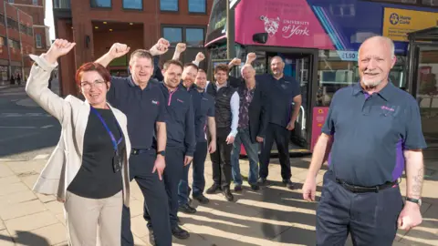 First Bus A group of people standing on a pavement in front of a pink bus. The bus features branding for “University of York” near the front door, along with prominent text indicating “Zero Emission Bus” across the top panel.