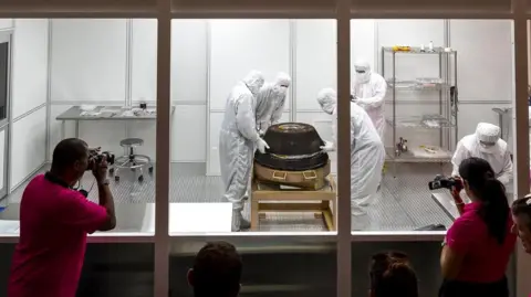 Nasa A Nasa team dressed in white suits and masks examining a space capsule in a cleanroom.