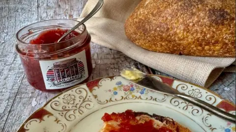 Single Variety Co A jar of jam on a wooden table with a label on it which says Jail Jars. Beside it there is an ornate china plate with a piece of jam toast on it and a butter knife resting on the side. Also on the table is a crusty loaf of bread laying on a burlap sack. 