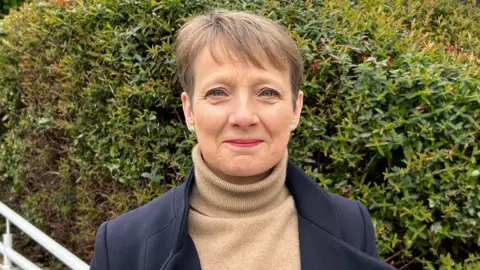 Clare Moody, looking towards the camera. She is wearing a blue coat and brown top, and is standing in front of some plants