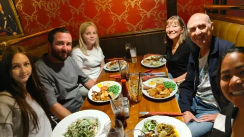 Steve Cervi A family group sit around a table in a booth at a restaurant with food and drinks laid out in front of them. On the left is a man with two teenager girls and on the right is an older woman, and older man and a young woman. 