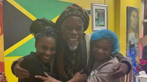 Mama Flo's Three people are standing closely together inside a brightly lit room, embracing each other in a group hug. The person in the center is wearing a colorful patterned outfit with a knitted hat featuring red, green, and yellow stripes. The two individuals on either side are dressed casually, one in a dark apron over clothing and the other in a light sweatshirt with a blue head covering. Behind them, a large Jamaican flag is displayed on a yellow wall, along with framed certificates and posters. A white chair and a table are visible to the side, and the floor is tiled in a light pattern.