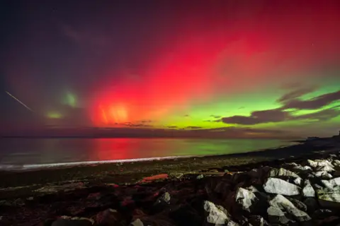 Mark Hetherington The beach in the foreground is rocky and the sea in front of it is calm. Above, the northern lights have illuminated the sky pink and green. On the left, there is a small bright streak indicating a meteor.