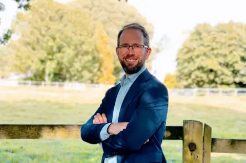 Conservative Party Conservative candidate Matthew Barber standing in front of a wooden fence, with a field in the background