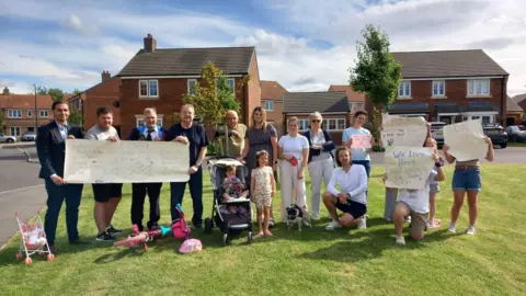 About a dozen people stand holding paper signs on a green in front of some houses. The signs read: "We live here" and "Don't destroy our woods".