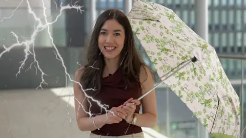 A woman with brown hair, wearing a brown vest top is holding a green and white umbrella. 