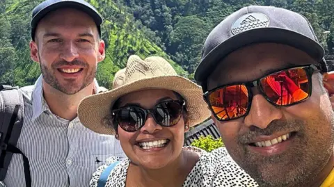 Three people in sunglasses and hats smile at the camera. Behind them are green mountains and trees