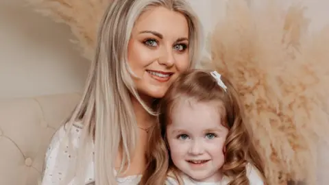 Hollie pictured on a cream chair with her mum Ann, the photo is professional. Hollie has long curly red/brown hair and is wearing a blue and white dress with a white cardigan. Ann has long blonde hair and is holding Hollie. 