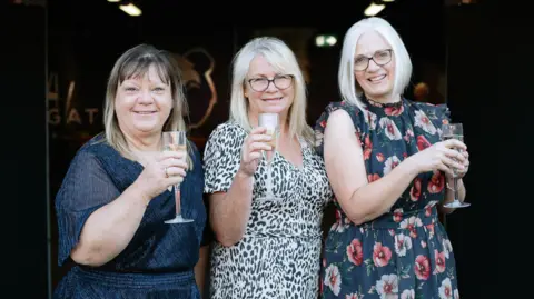 Kathy, Sarah and Debbie all holding a glass of champagne, smiling at the camera 