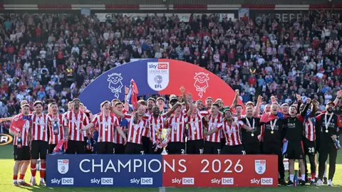 The Lincoln City players in red and white striped shirts and black shorts celebrate on the pitch with the League One champions trophy