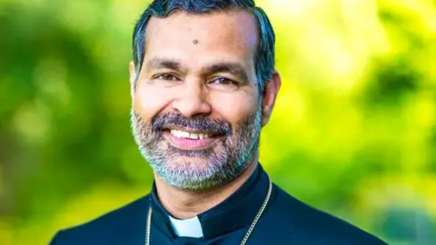 Church of England Right Reverend Dr John Perumbalath who has a grey beard smiles in front of a blurry green background