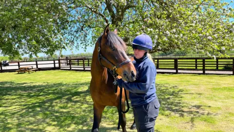 TOM JACKSON/BBC A horse rider standing next to a light brown horse. The woman is wearing a riding helmet and holding the animal by a reign. They are standing in a gated paddock with grass and trees. 