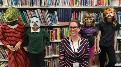 YorKAOAS The four children from the art project are stood in a library with their masks on, and with a member of library staff who is a woman in a navy blue, red and white striped dress and glasses.