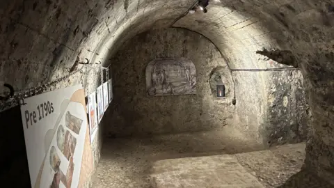 Emma Craig/BBC A basement made of yellow stone. It has an arched roof. At the back is a line drawing showing people and a bucket. There are information boards on the right hand side. 