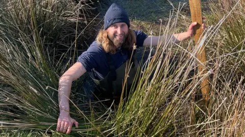 RSPB Dan Cropper amongst some reeds. He is wearing a blue hat and has a beard and long hair.