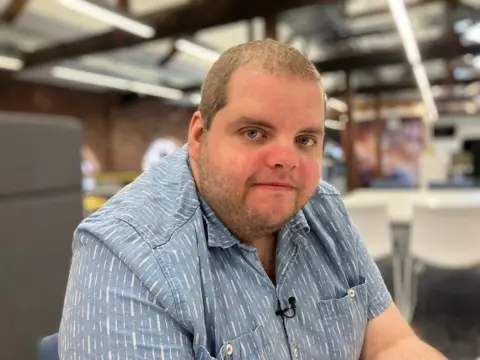 A young man wearing a blue shirt looks at the camera. He has light hair and stubble.