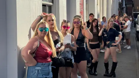 A group of people stand in a long queue outside a building on a street.
