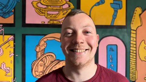 A man smiling at the camera with very short hair and wearing a red shirt
