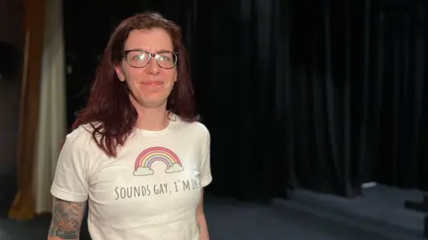 Kaye smiles at the camera with a white t-shirt on with a rainbow on it and text saying 'Sounds Gay, I'm In'. She has dark red hair which comes down past her shoulders and is wearing glasses. Behind is are the black stage curtains.