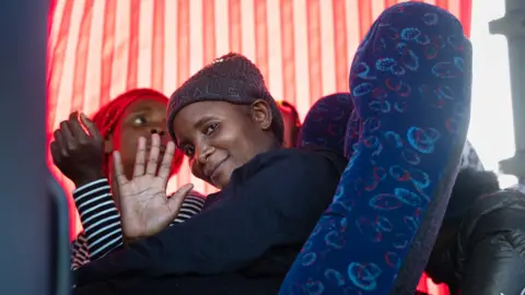 A woman in a grey hat sitting in a coach smiles and waves from the window at the people outside. 
