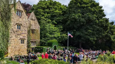 About 200 hundred people gather in the grassy gardens outside the tall stone building. There is bunting hung across the manicured hedges between the crowds and the manor. A flag pole in the middle of the grounds is flying an red, white and blue American flag. There are tall, lush green trees in the background.