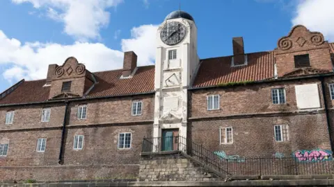 The Keelman's Hospital is a two-storey brown brick building with white window panes and a white clock tower with a small dome in the middle. Parts of the building are covered in pink and blue graffiti. 